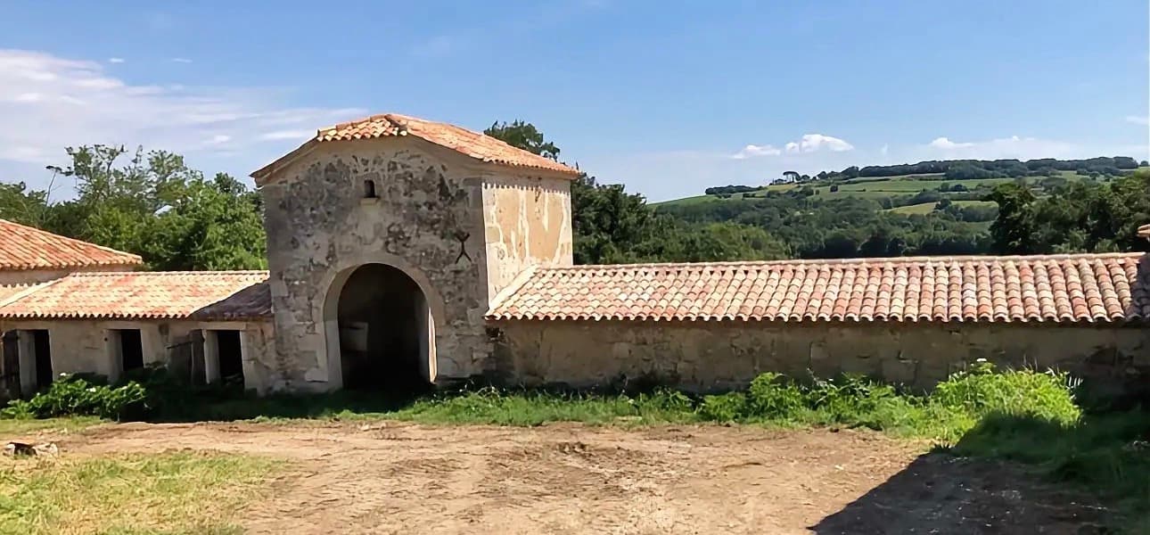 Bâtiment rural en pierre avec entrée voûtée et toit de tuiles devant des collines verdoyantes.